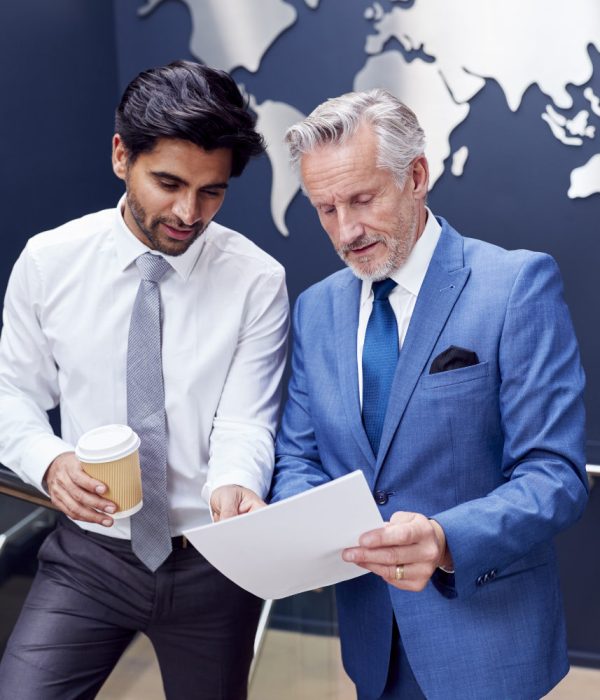 Male Colleagues Meeting On Stairs Of Office Discussing Document With World Map In Background